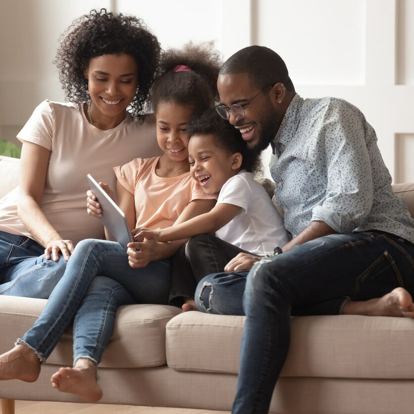 Happy african family having fun with device at home, black parents and little children using digital tablet looking at screen sit on sofa together, cute kids hold computer laugh watch cartoons online